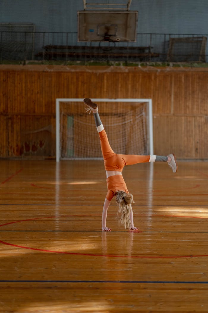 hero-img-02 A young girl in orange attire performs a cartwheel on a gymnasium floor, showcasing her gymnastics skills.