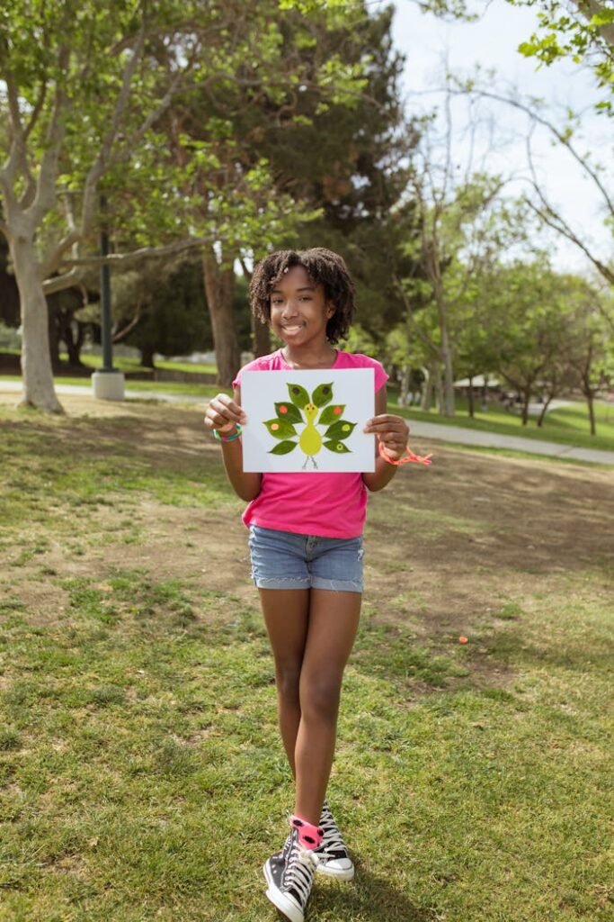 A cheerful young girl holds a colorful leaf artwork outdoors in a sunny park.